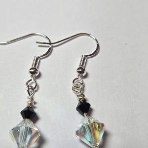 May include: A pair of silver-toned earrings with a fish hook design. Each earring features a black, faceted bead above a larger, clear, faceted crystal. The crystals reflect multiple colors. The earrings are displayed against a white background.
