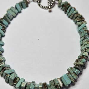 May include: A turquoise chip bead bracelet with a silver-colored toggle clasp. The bracelet features irregular, rectangular turquoise beads with brown matrix patterns. The clasp is a simple, twisted design.