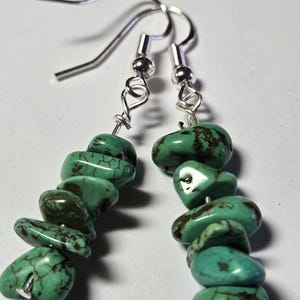 May include: A pair of turquoise stone earrings. Each earring features a series of polished, irregular turquoise stones strung together. The stones are a vibrant turquoise color with brown veining. The earrings have silver-colored ear wires.