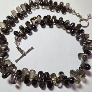 May include: A necklace featuring teardrop-shaped smoky quartz beads in varying shades of brown and gray. The beads are strung together with a silver-colored clasp and chain. The necklace is displayed on a white surface.