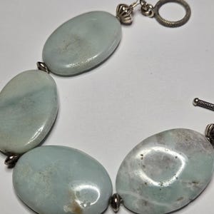 May include: A bracelet featuring large, oval-shaped, pale blue-green stones. The stones are connected by small silver-toned metal links and a toggle clasp. The bracelet has a polished, smooth surface and a natural, organic aesthetic.