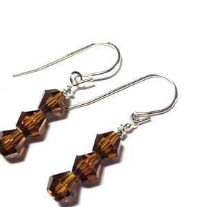 May include: A pair of dangle earrings featuring three faceted, amber-colored beads strung on a silver-tone hook. The beads have a geometric shape and reflect light, creating a sparkling effect. The earrings are displayed against a white background.