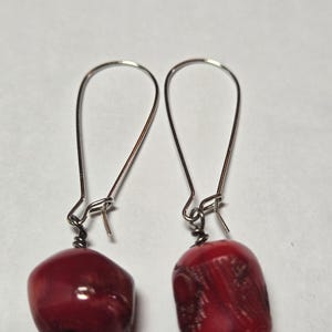 May include: A pair of dangle earrings featuring large, polished red coral beads. The beads are suspended from silver-toned, curved ear wires. The earrings have a simple, elegant design.