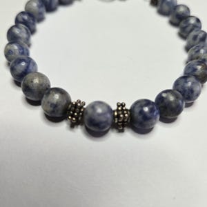 May include: A beaded bracelet featuring round, blue-grey sodalite stones. The bracelet has a silver-toned clasp and decorative metal accents between some of the stones. The stones have a speckled appearance.