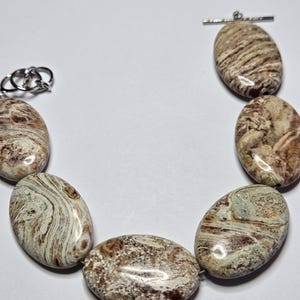May include: A necklace featuring large, oval-shaped beads in shades of brown, cream, and tan. The beads have a natural, marbled appearance and are strung together with a silver-colored clasp and toggle closure. The jewelry is displayed against a white background.
