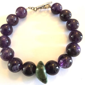 May include: A beaded bracelet featuring large, round, deep purple amethyst stones. The bracelet includes a single, elongated, green stone and a silver-colored clasp. The amethyst beads have a polished finish.