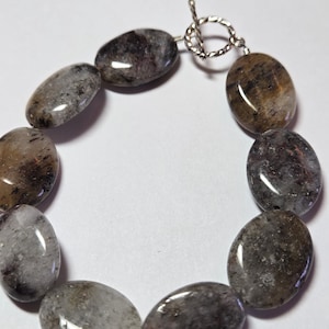 May include: A bracelet featuring oval-shaped, polished gemstones in shades of gray, brown, and clear. The stones are strung together with a silver-colored toggle clasp. The bracelet is displayed on a white background.