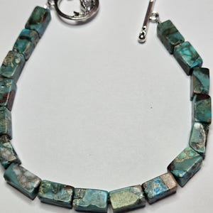 May include: A turquoise and brown stone bracelet with a silver toggle clasp. The bracelet features rectangular beads with a mix of blue, green, and brown hues. The clasp is a silver toggle bar and a decorative silver ring.
