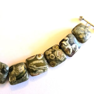 May include: A close-up of a necklace featuring square-shaped beads. The beads are primarily green with brown and white patterns. The necklace has a silver-colored clasp. The beads are approximately 0.75 inches square.