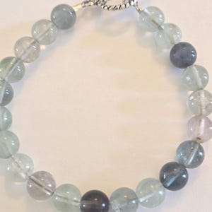 May include: A beaded bracelet featuring a variety of translucent, round beads in shades of green, gray, and purple. The bracelet is secured with a silver-colored toggle clasp.