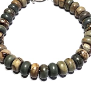 May include: A beaded necklace featuring a variety of earth-toned beads in shades of brown, green, and tan. The beads are strung together with a silver-colored clasp. The necklace is displayed against a white background.