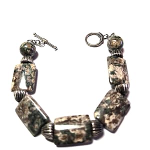 May include: A bracelet featuring rectangular and round beads in shades of green, brown, and cream. The beads are strung together with silver-colored metal accents and a toggle clasp. The bracelet is displayed against a white background.