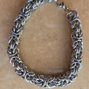 May include: A silver-colored Byzantine chain bracelet. The bracelet is made of interlocking metal rings, creating a textured and detailed design. It features a lobster clasp closure for secure wear. The jewelry is displayed on a light brown surface.