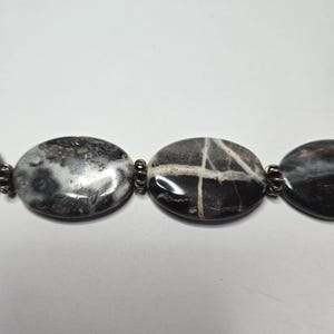 May include: Close-up of a necklace featuring large, oval-shaped beads. The beads are primarily black and gray with unique patterns and textures. The beads are connected by small, silver-colored metal links. The necklace is set against a white background.