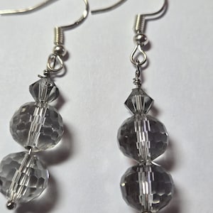 May include: A pair of dangle earrings featuring faceted gray beads. Each earring has a silver-colored fish hook and is strung with two large, round beads and one smaller, diamond-shaped bead. The beads have a reflective, crystal-like appearance.
