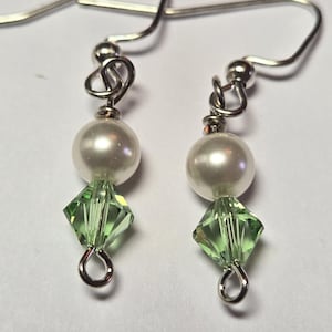 May include: A pair of dangle earrings featuring a white pearl bead and a green, diamond-shaped crystal. The earrings have silver-colored hooks and connecting wires, creating a delicate and elegant design.