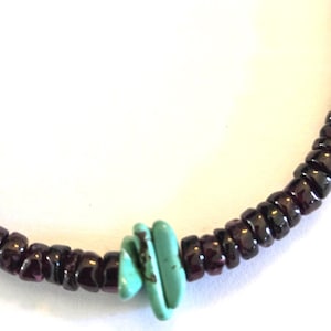 May include: Close-up of a beaded bracelet. The bracelet features small, dark red beads and a cluster of three turquoise-colored, stacked beads. The beads are strung together, creating a curved shape against a white background.