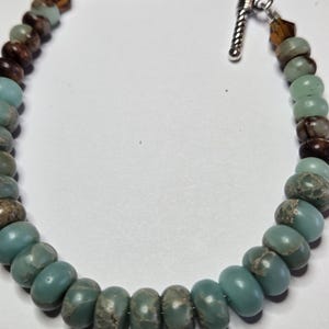 May include: A beaded bracelet featuring turquoise, brown, and clear beads. The bracelet has a silver toggle clasp. The beads are oval-shaped and strung together. The bracelet is on a white background.