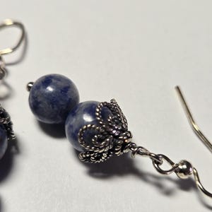 May include: Close-up of a pair of earrings featuring dark blue, spherical beads. The beads are accented with ornate, silver-colored metalwork. The earrings have a hook closure and are set against a white background.