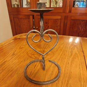 May include: A wrought iron candle holder with a heart-shaped design. The candle holder has a circular base, a twisted stem, and a small dish to hold a candle. The metal is a dark grey colour.