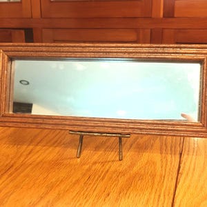 May include: A rectangular wooden framed mirror with a light brown finish. The mirror is supported by a small, gold-colored metal stand. The mirror is displayed on a wooden table.