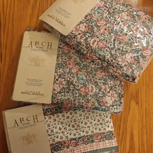 May include: Three packages of floral print bed sheets from the Arch Home Collection. The sheets feature a pattern of pink, blue, and green flowers on a dark background. The packaging includes the text "100% Combed Cotton Luxury Percale".