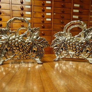 May include: Two silver-colored napkin holders. Each holder features a grape cluster design with leaves and a handle. The holders are placed on a wooden surface, with a wooden cabinet in the background.