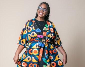 Op de afbeelding: Een kleurrijke kimono-stijl jurk met een levendig geometrisch en verenpatroon in blauw, geel, oranje en zwart. De jurk heeft een wikkelontwerp met een bijpassende riem. De mouwen zijn ellebooglengte.