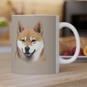 Könnte beinhalten: Eine weiße Keramiktasse mit braunem Hintergrund, auf der eine detaillierte Illustration eines Shiba Inu Hundes mit braunem und weißem Fell zu sehen ist. Der Hund schaut den Betrachter direkt an und hat einen freundlichen Ausdruck.