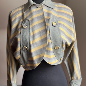 Könnte beinhalten: Eine Vintage-Jacke mit horizontalen grauen und gelben Streifen. Die Jacke hat einen Kragen, Knöpfe und dekorative Einsätze mit passenden Knöpfen. Die Ärmel sind umgeschlagen, und die Jacke wird auf einer schwarzen Schaufensterpuppe präsentiert.