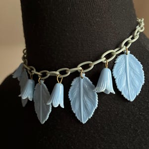 May include: A light blue necklace with leaf and bell-shaped charms. The charms are suspended from a gray chain. The necklace is displayed on a black mannequin. The necklace is a fashion accessory.