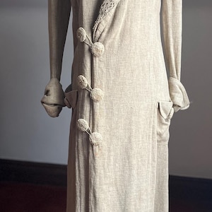 May include: A vintage beige dress with a long, flowing silhouette. The dress features a decorative lace collar, long sleeves, and unique button closures. Two pockets are visible on the front. The dress is displayed on a mannequin.