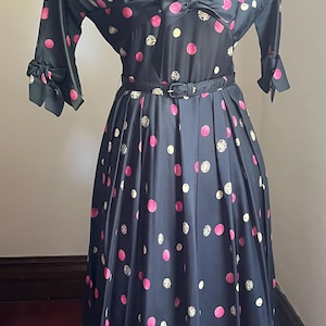 Puede incluir: Vestido vintage azul marino con lunares rosas y blancos. Cuello blanco, corpiño entallado, detalle de lazo y falda acampanada. Mangas largas con detalles en los puños y cinturón a juego.