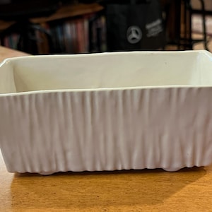 May include: A rectangular, off-white ceramic planter with a textured, vertical ribbed design. The planter has a slightly flared shape and is suitable for indoor or outdoor use. The neutral color complements various decor styles.