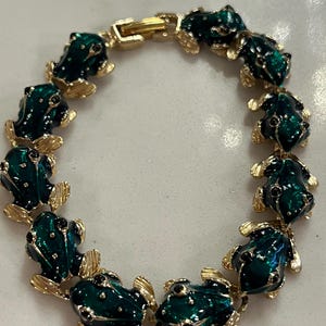 May include: A gold-toned bracelet featuring a series of green frog charms. Each frog has black accents and is connected by gold-colored links. The bracelet has a secure clasp.