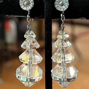 May include: A pair of clear, faceted crystal drop earrings featuring a tiered, geometric design. Each earring consists of a circular stud top connected by a silver-toned metal link to three stacked, graduated crystal beads that taper to a point.