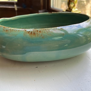 Puede incluir: Un cuenco de cerámica turquesa con acabado brillante. El cuenco tiene forma redondeada con un borde ligeramente irregular y una banda marrón alrededor del borde superior. El cuenco está sobre una superficie blanca.