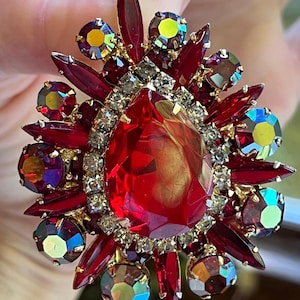 May include: A large, ornate brooch with a central teardrop-shaped red gemstone, surrounded by smaller red and iridescent stones. The brooch features a gold-toned metal setting and is encrusted with clear crystals. The design is symmetrical and eye-catching.