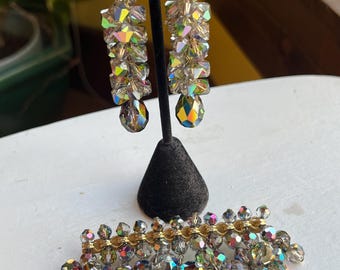 May include: A vintage jewelry set featuring a pair of dangling earrings and a brooch. The earrings and brooch are adorned with iridescent, faceted crystals in shades of gray, gold, and rainbow hues. The earrings are suspended from gold-toned metal.