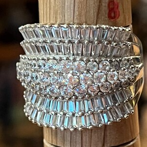 Puede incluir: Un anillo de tono plateado con tres filas horizontales de piedras transparentes facetadas. Las filas superior e inferior consisten en piedras rectangulares de corte baguette, mientras que la fila central contiene piedras redondas de corte brillante. El anillo está colocado en un mandril de madera marcado con el número 8.
