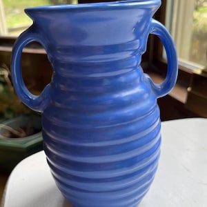May include: A blue ceramic vase with two handles. The vase has a spiral design and a wide opening. The vase is sitting on a white surface. The vase is a medium shade of blue.