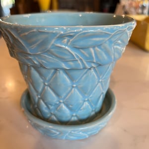 Puede incluir: Una maceta de cerámica azul claro con un platillo a juego. La maceta tiene un patrón de diamantes acolchados y diseños de hojas. El platillo tiene un diseño de hojas a juego. Un artículo decorativo para plantas.