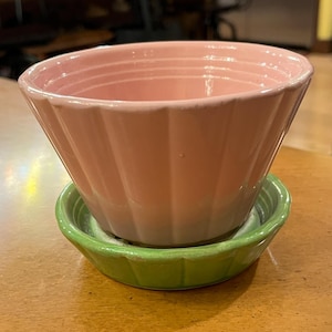 May include: A pink ceramic bowl with a fluted design sits atop a small, green saucer. The bowl has a wide, open top and a slightly tapered shape. The saucer is round with a similar fluted edge. The items are on a wooden surface.