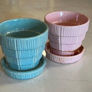 May include: Two ceramic plant pots, one light blue and one pink, each with a matching saucer. The pots have a textured, basket-weave design. The pots are empty and ready for planting.