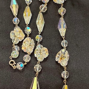 Puede incluir: Un conjunto de joyas vintage con un collar y pendientes a juego. El collar tiene tres hilos de cuentas claras y facetadas y detalles florales. Los pendientes son redondos con detalles de cristal transparente. El conjunto está sobre un fondo negro.