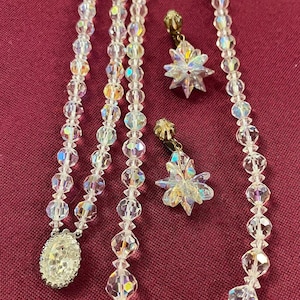 May include: A vintage jewelry set featuring a necklace and earrings. The necklace has a large oval pendant and is strung with clear, faceted beads. The earrings are star-shaped with similar beads. The set is displayed on a burgundy surface.