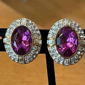 May include: A pair of oval-shaped earrings featuring a large, faceted purple center stone set in a gold-tone frame. The border is encrusted with small, clear round crystals and accented by four rectangular clear stones at the cardinal points.