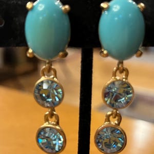 May include: A pair of gold-toned drop earrings. Each earring features a large, oval, turquoise-colored stone at the top, with two smaller, round, light blue crystal stones dangling below. The earrings are displayed against a black background.