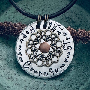 Könnte beinhalten: Silberne Halskette mit einem runden Anhänger, der mit einem dekorativen Filigranmuster verziert ist und die Worte "Hope", "Courage", "My Strength" und "My Best" am Rand eingraviert sind.