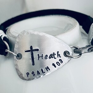 Puede incluir: Un brazalete de cuero negro con un dije de púa de guitarra plateado. El dije tiene una cruz y el texto "Heath Psalm 103" grabado.